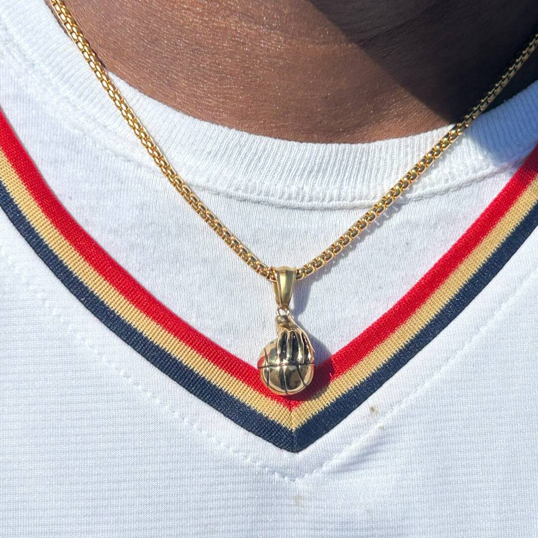 Gold plated hand palming basketball pendant on a 3mm box chain necklace worn over a white shirt with red, navy, and yellow stripes.