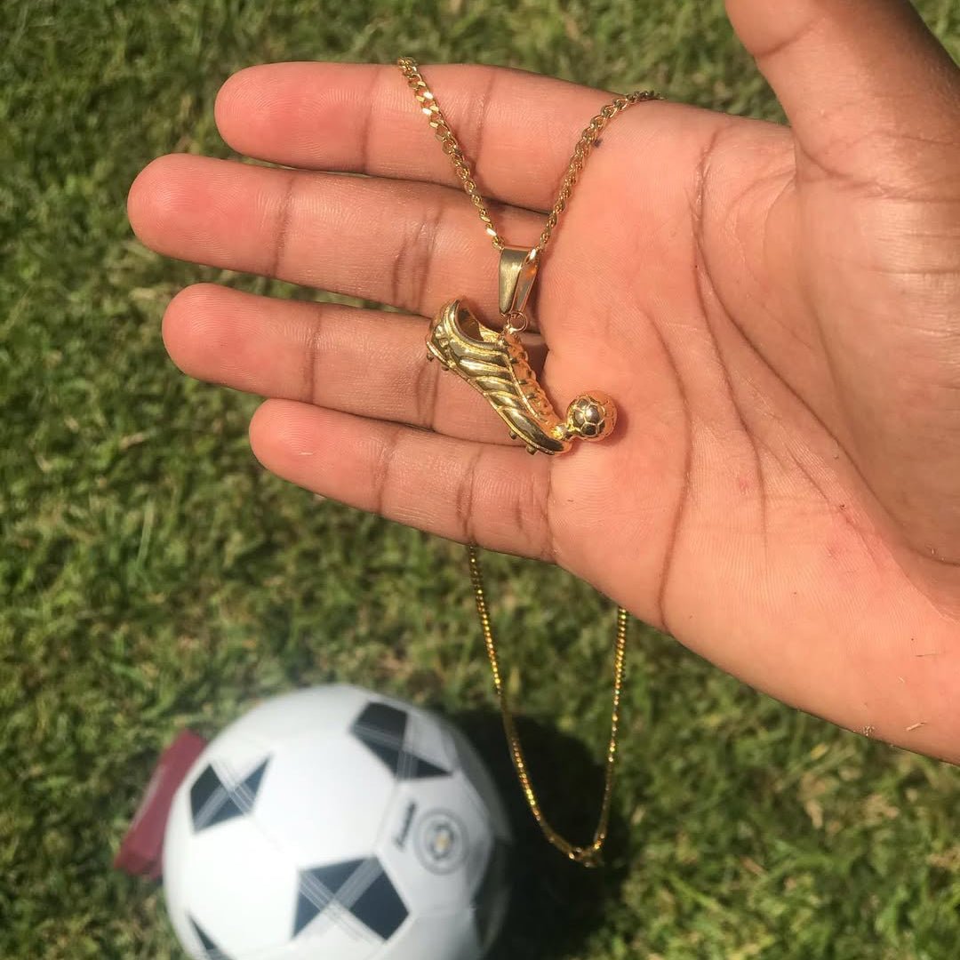 Gold-plated soccer cleat pendant with ball on Cuban link chain held in hand outdoors with soccer ball in background