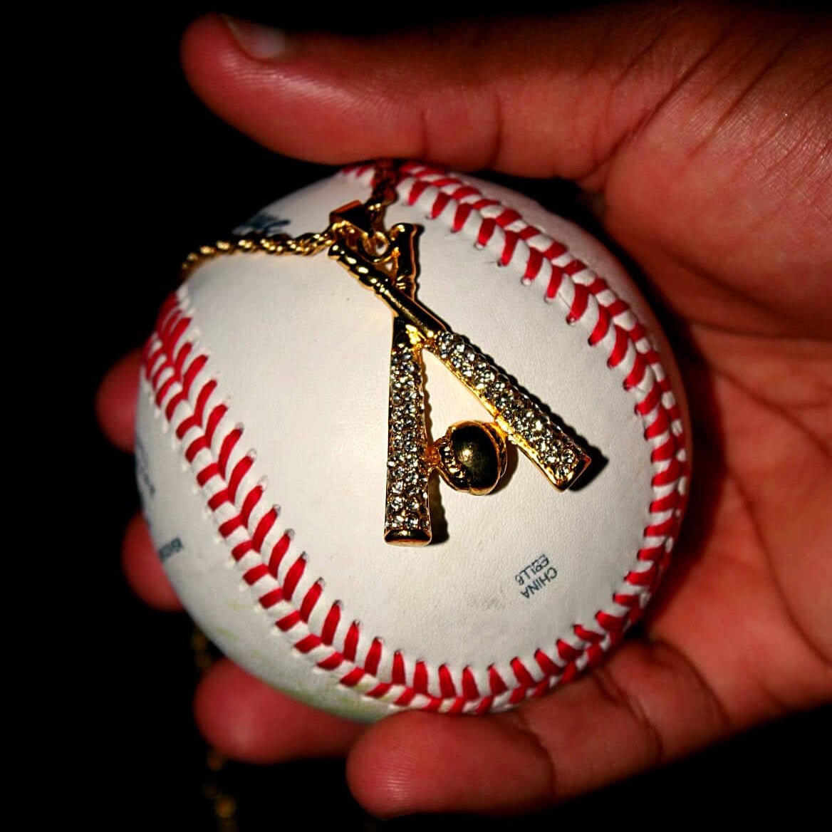Gold studded baseball bats pendant and chain necklace held on a white baseball with red stitching