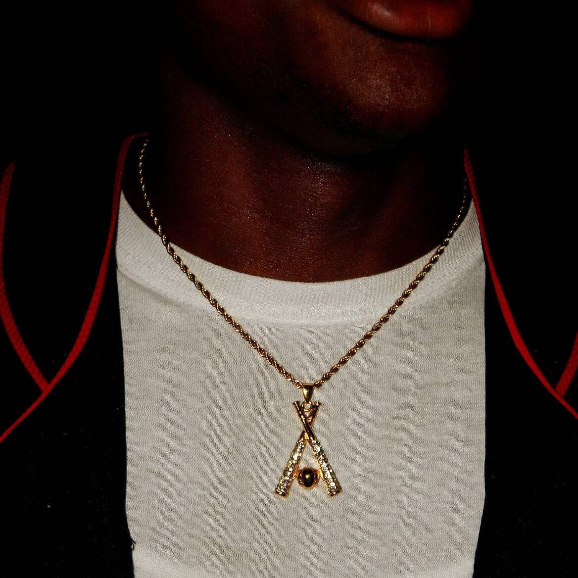 Close-up of a studded baseball bats pendant on a gold rope chain necklace worn over a white shirt