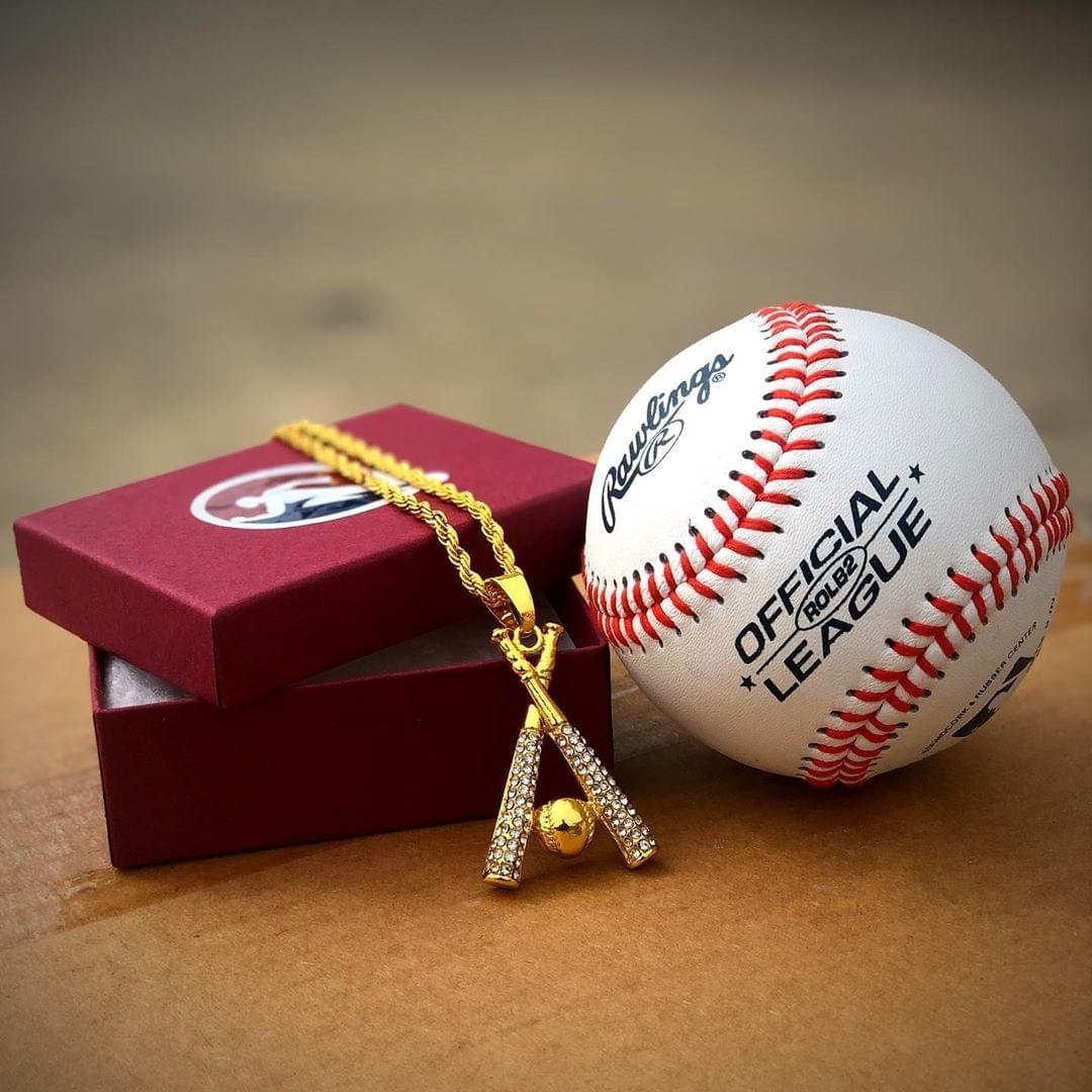 Gold studded baseball bats pendant necklace with cubic zirconia stones and rope chain next to baseball and red gift box
