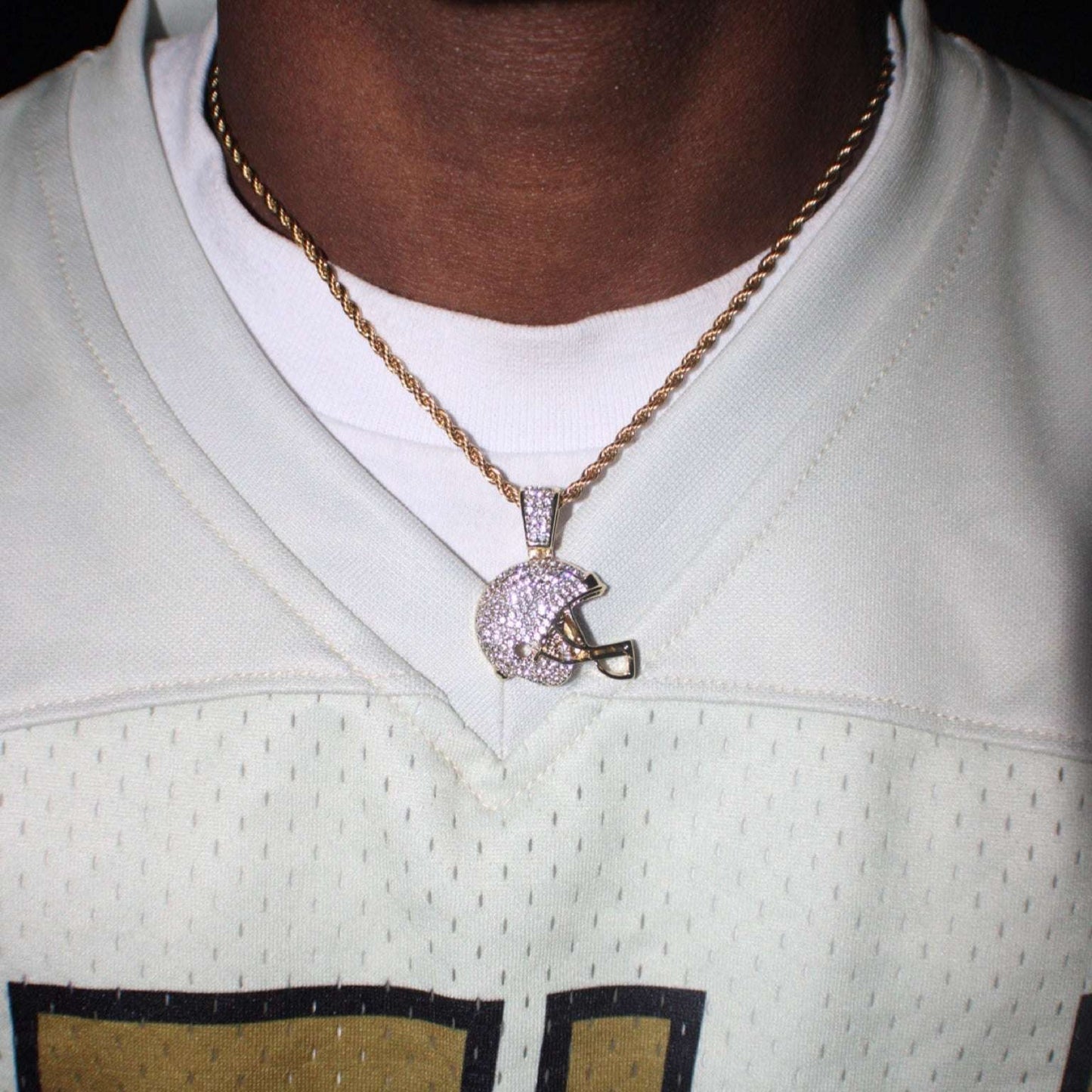 Close-up of a studded football helmet pendant on a gold rope chain necklace worn over a white football jersey