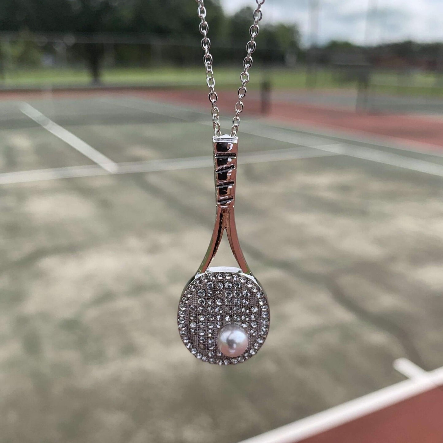 Studded tennis racket pendant necklace with cubic zirconia stones and pearl on silver chain displayed on tennis court background