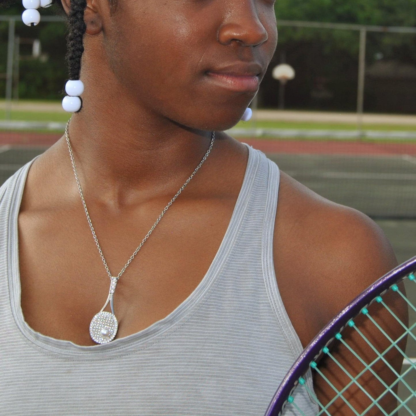 Close-up of a studded tennis racket pendant necklace with cubic zirconia and pearl tennis ball worn by a woman on a chain outdoors near a tennis court