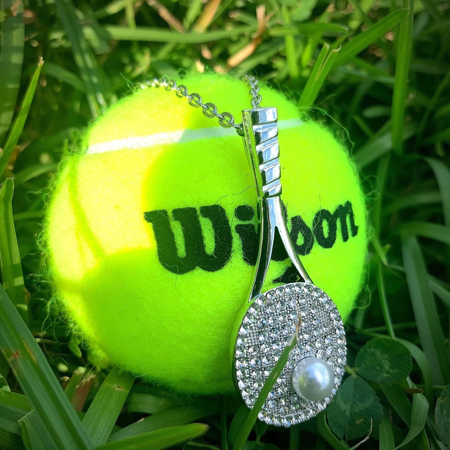 Studded tennis racket pendant necklace with cubic zirconia and pearl on silver chain displayed on tennis ball in grass