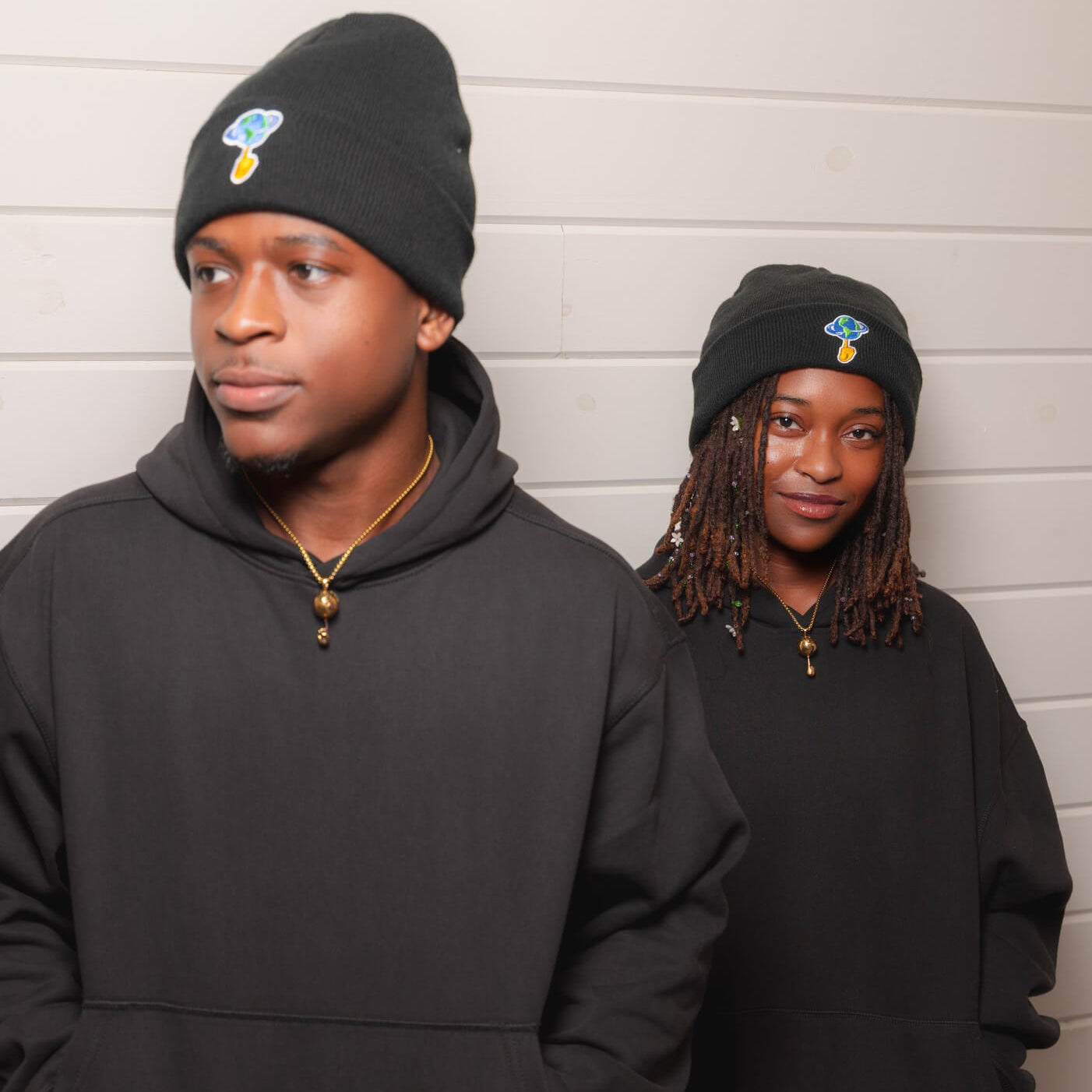 Two models wearing matching black hoodies and beanie caps with a small blue and gold graphic logo on the front, standing against a white paneled wall.