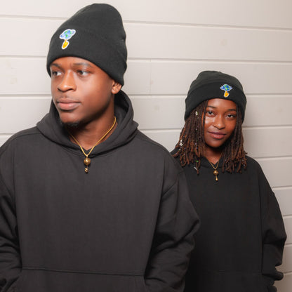 Two models wearing matching black hoodies and beanie caps with a small blue and gold graphic logo on the front, standing against a white paneled wall.