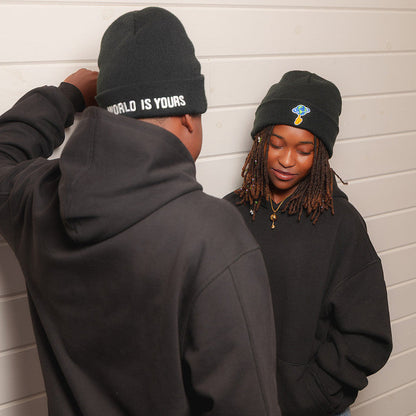 Two people wearing black beanie hats and black hoodies standing against a white paneled wall. The person on the left is shown from the back, wearing a black beanie with white text that reads 'WORLD IS YOURS'.