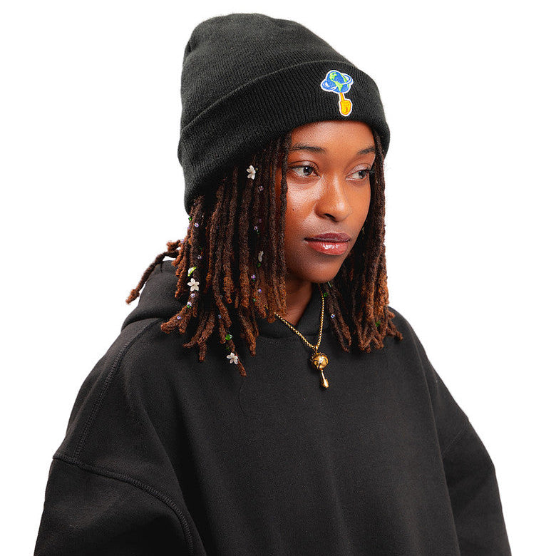 A person wearing a black knit beanie with a small colorful embroidered cloud patch and a black oversized hoodie, photographed against a plain white background.