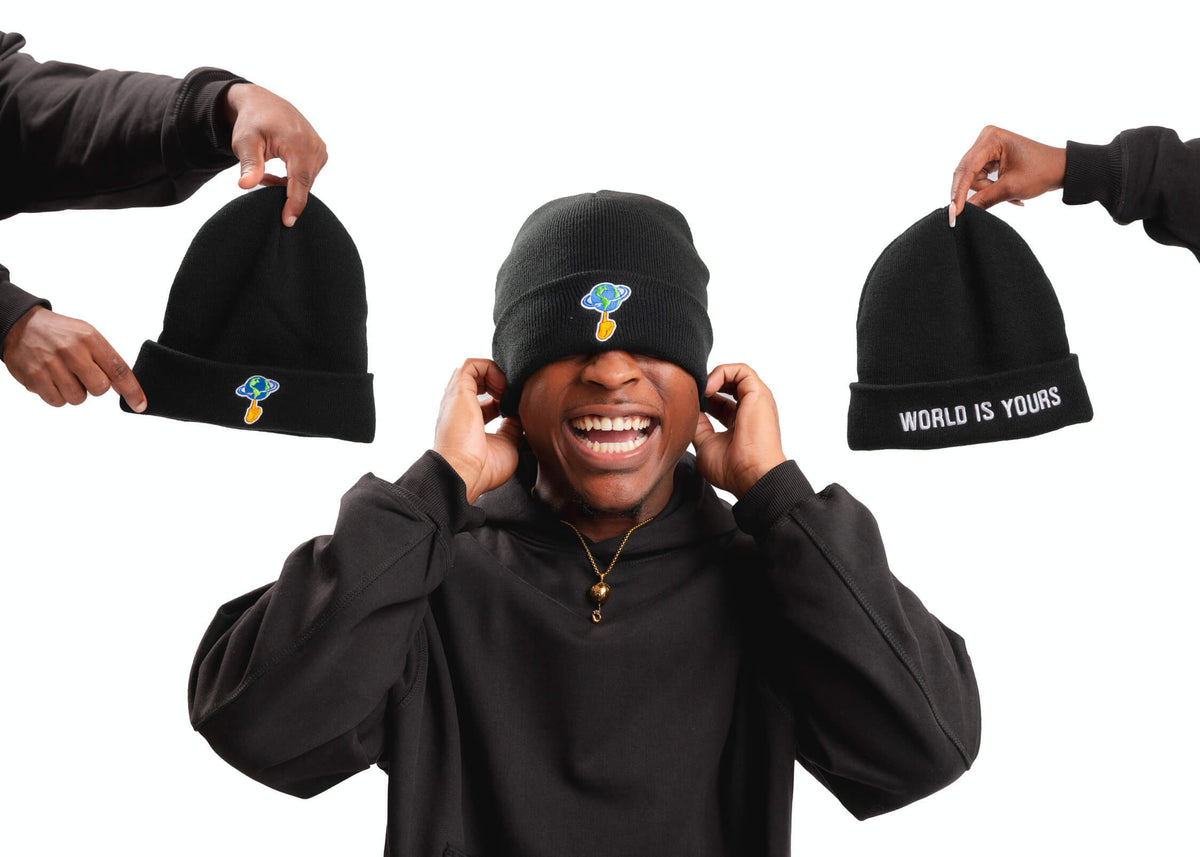A person in a black hoodie wears a black beanie with a small colorful mushroom graphic on the front. Two other hands at the edges hold up matching black beanies, one with white text reading 'WORLD IS YOURS' and the other with the small mushroom graphic.
