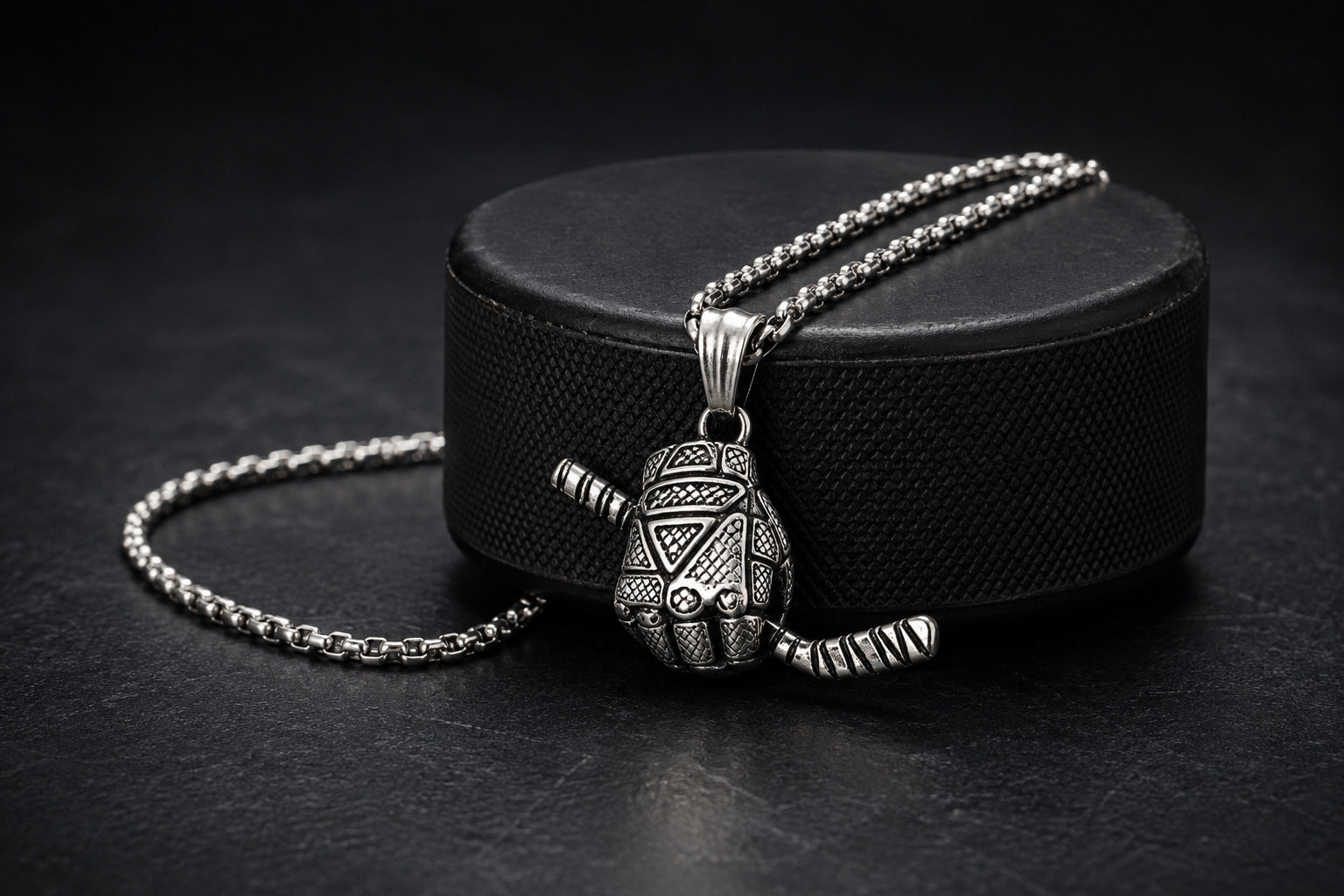A silver-toned pendant necklace with a detailed hockey glove pendant, displayed on a black hockey puck against a dark background.